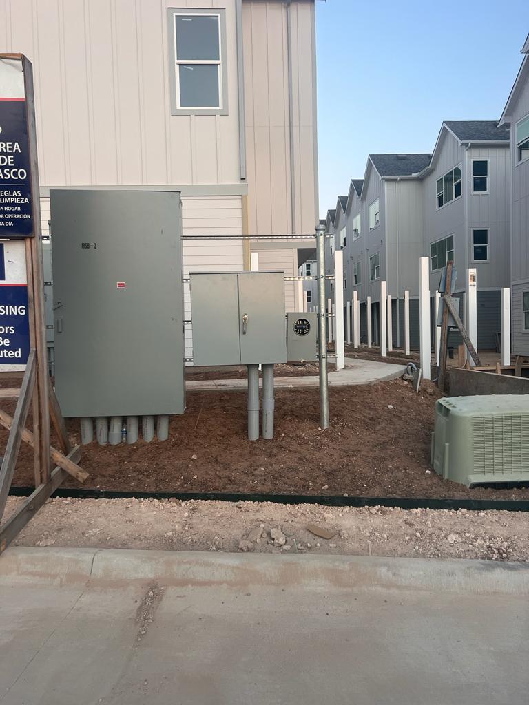 Multiple electrical panels and a meter installed on a pole at a construction site with newly built houses in the background.