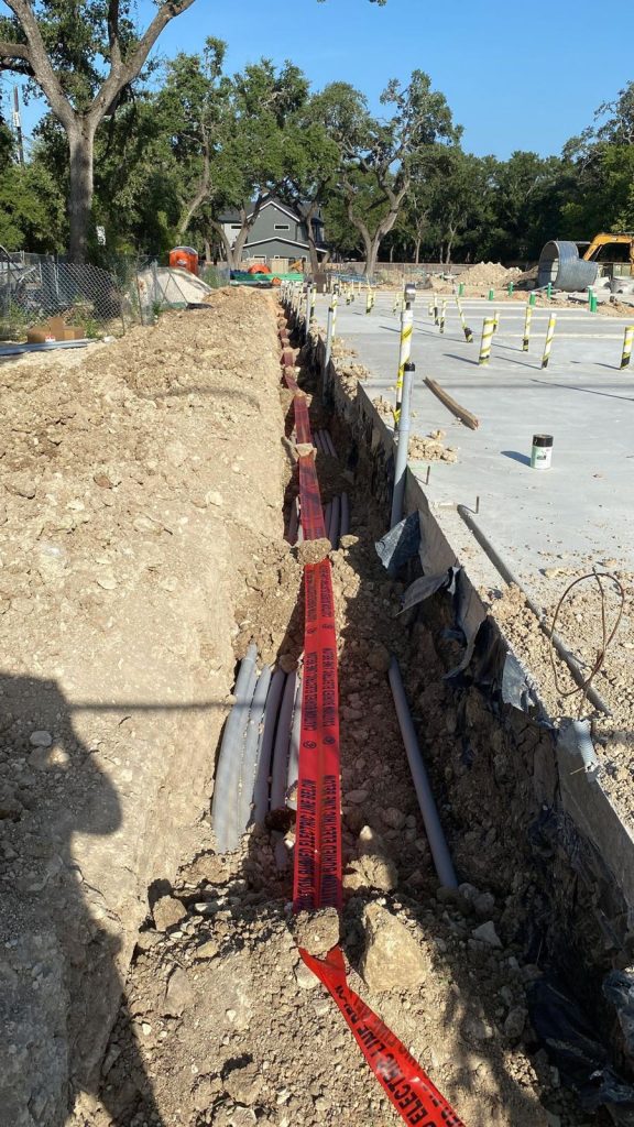A trench dug at a construction site with electrical piping and bright red warning tape.