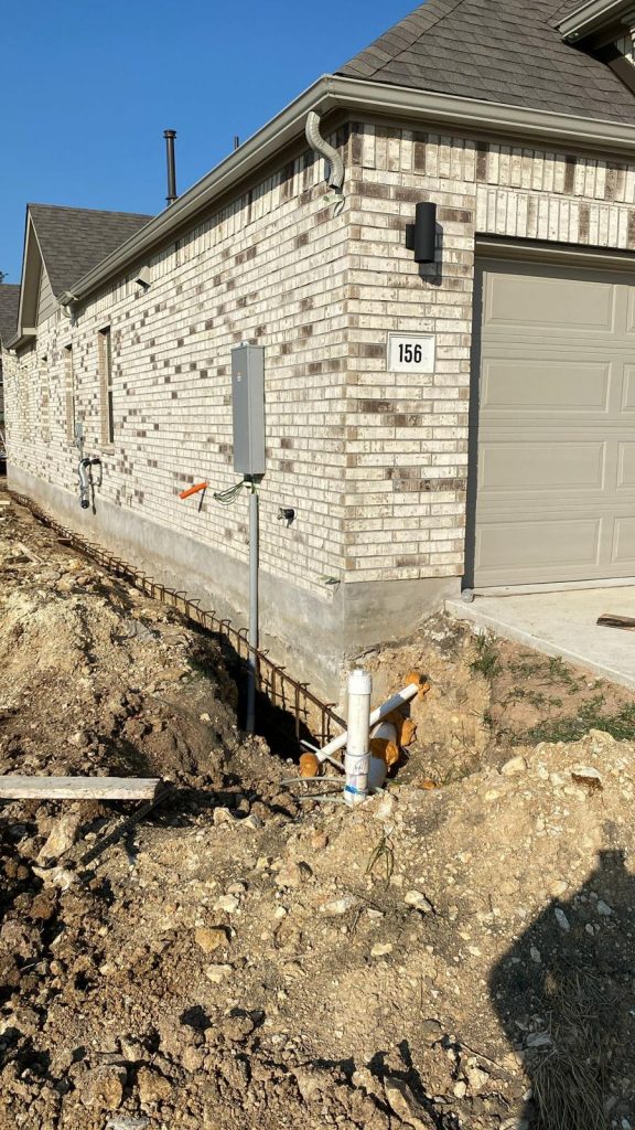 Newly constructed brick house with a trench that runs along the side of the house, exposing pipes and electrical conduits. A meter box is mounted on the brick wall.