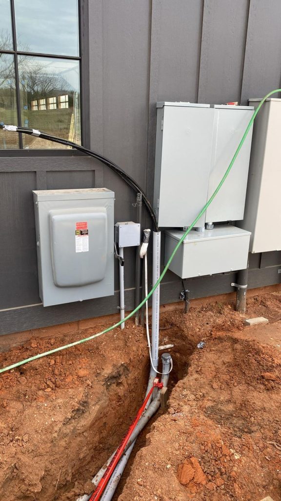 Electrical conduit and underground wiring, connected to a gray meter box and electrical panels mounted on a building's exterior wall.