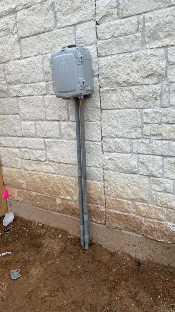 Gray electrical meter box mounted on a brick wall with two metal conduits extending into the ground.