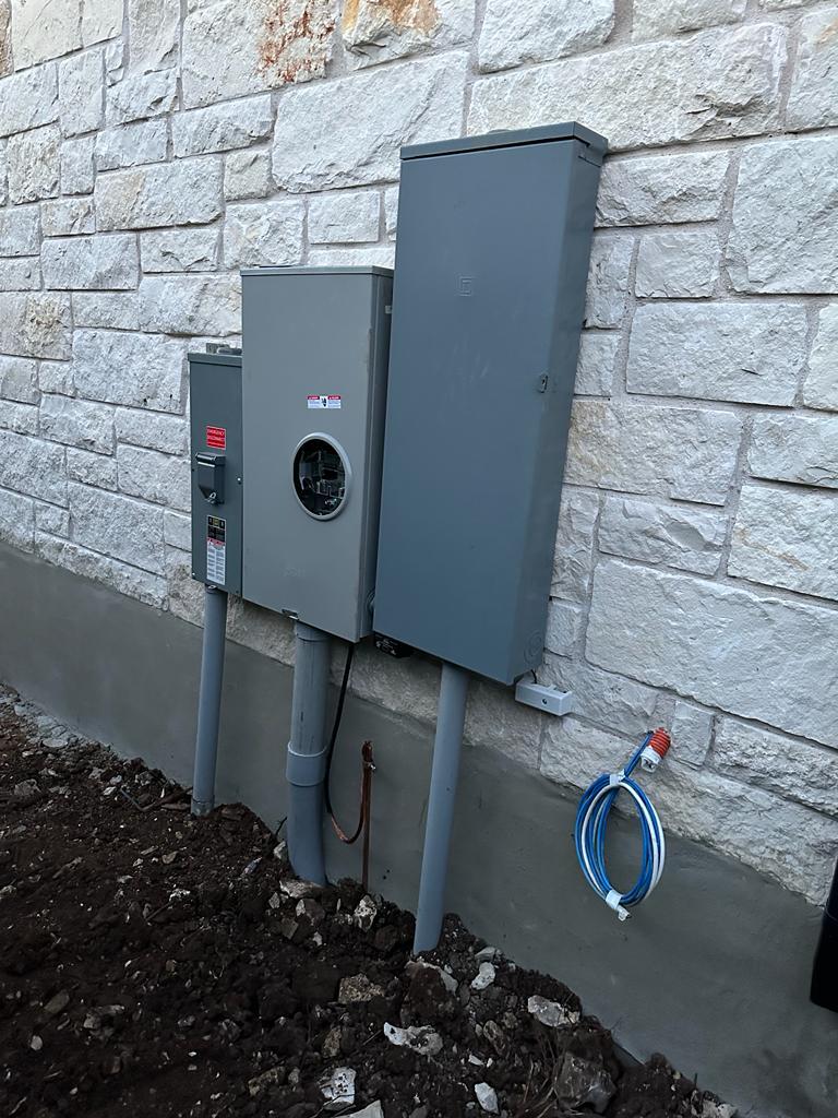 Three gray electrical panels, including a meter box, mounted on a stone wall with conduit and wiring connections.