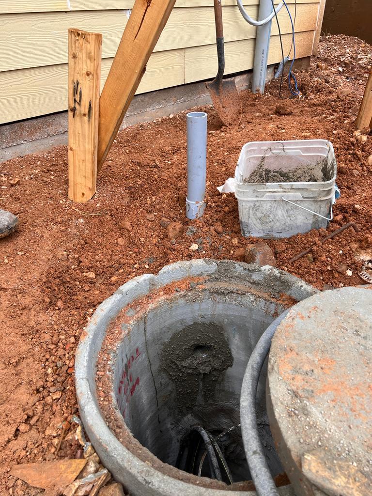Open concrete manhole revealing pipes and electrical conduit. Some construction tools and materials are visible in the background.