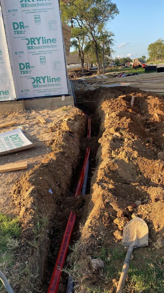 Trench dug in the ground with grey conduits laid inside and red tape.