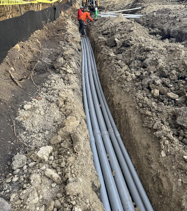 Two construction workers installing underground piping and an excavator next to them.